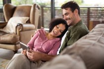 Heureux couple diversifié assis sur le canapé dans le salon en utilisant la tablette. passer du temps à la maison dans un appartement moderne. — Photo de stock