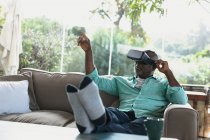 Uomo anziano afroamericano seduto sul divano e con auricolare vr nel soggiorno moderno. stile di vita di pensione, trascorrere del tempo da solo a casa. — Foto stock