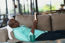 Relaxante homem afro-americano sênior deitado no sofá e lendo livro na sala de estar moderna. estilo de vida aposentadoria, passar o tempo sozinho em casa. — Fotografia de Stock