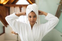 Портрет усміхненої змішаної раси жінки у ванній кімнаті, що зав'язує рушник на голові, дивлячись на камеру. домашній спосіб життя, насолоджуючись доглядом за дозвіллям вдома . — стокове фото