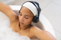 Femme de race mixte dans la salle de bain relaxant dans la baignoire avec casque, les yeux fermés. mode de vie domestique, profiter de loisirs d'auto-soins à la maison. — Photo de stock