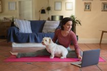 Lächelnde Kaukasierin im Wohnzimmer mit ihrem Hund, Yoga praktizierend, mit Laptop. häuslicher Lebensstil, Freizeit zu Hause genießen. — Stockfoto