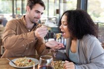 Happy diverse couple in living room sitting at table eating dinner together. spending time off at home in modern apartment. — Stock Photo