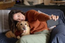 Усміхнена біла жінка у вітальні, сидячи на дивані, обіймає свого домашнього собаку, приймаючи селфі. домашній спосіб життя, насолоджуючись дозвіллям вдома . — стокове фото