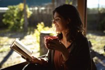 Donna mista lettura libro corsa e bere caffè in giardino soleggiato. stile di vita sano, godendo del tempo libero a casa. — Foto stock