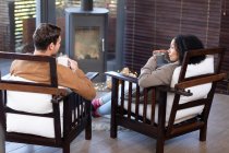 Heureux couple diversifié dans le salon assis près de la cheminée tenant des tasses et buvant du café. passer du temps à la maison. . — Photo de stock