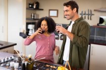 Felice coppia diversificata in cucina preparare il cibo insieme bere vino. trascorrendo del tempo a casa in un appartamento moderno. — Foto stock
