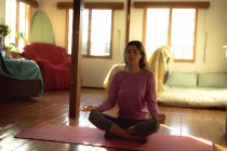 Mujer de raza mixta practicando yoga, sentada meditando en la soleada sala de estar. estilo de vida saludable, disfrutando del tiempo libre en casa. - foto de stock