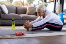 Mujer caucásica mayor en la sala de estar sentada en el suelo y haciendo ejercicio. estilo de vida de jubilación, pasar tiempo solo en casa. - foto de stock