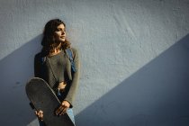Смешанная расовая женщина держит скейтборд в солнечный день на улице. здоровый образ жизни, наслаждаясь отдыхом на открытом воздухе. — стоковое фото