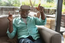 Felice uomo afroamericano anziano seduto e utilizzando auricolare vr nel soggiorno moderno. stile di vita di pensione, trascorrere del tempo da solo a casa. — Foto stock