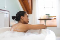 Femme mixte souriante dans la salle de bain relaxant dans la baignoire. mode de vie domestique, profiter de loisirs d'auto-soins à la maison. — Photo de stock