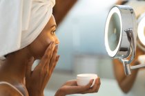 Змішана расова жінка у ванній, що наносить крем для догляду за шкірою, дивлячись у дзеркало. домашній спосіб життя, насолоджуючись доглядом за дозвіллям вдома . — стокове фото