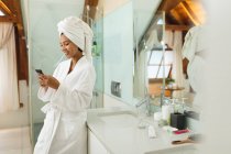 Mixed race woman in bathroom using smartphone and brushing her teeth. domestic lifestyle, enjoying self care leisure time at home. — Stock Photo