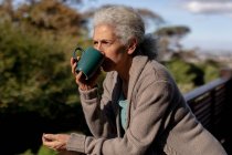 Détente femme caucasienne âgée sur le balcon debout et boire du café. mode de vie à la retraite, passer du temps seul à la maison. — Photo de stock