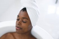 Улыбающаяся африканская американка в ванной комнате расслабляется в ванной с закрытыми глазами. домашний образ жизни, наслаждаясь отдыхом на дому. — стоковое фото