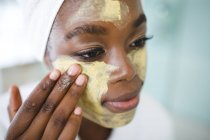 Smiling african american woman in bathroom applying beauty face mask. domestic lifestyle, enjoying self care leisure time at home. — Stock Photo