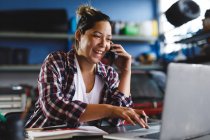 Smiling mixed race female car mechanic using smartphone and laptop in workshop. independent business owner at car servicing garage. — Stock Photo