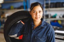 Mécanicien automobile mixte féminin en salopette, tenant un pneu, regardant la caméra. propriétaire d'entreprise indépendant au garage d'entretien de voiture. — Photo de stock