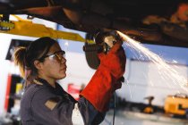Mixed race female car mechanic wearing overalls, using angle grinder. independent business owner at car servicing garage. — Stock Photo
