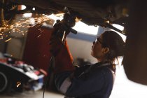 Corrida mista feminino carro mecânico vestindo macacão, usando moedor de ângulo. proprietário de empresa independente na garagem de manutenção de carro. — Fotografia de Stock