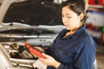 Mécanicien automobile mixte féminin portant des combinaisons, inspectant la voiture et utilisant une tablette. propriétaire d'entreprise indépendant au garage d'entretien de voiture. — Photo de stock