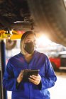 Mixed race female car mechanic wearing face mask and overalls, making notes on clipboard. independent business owner at car servicing garage during covid 19 pandemic. — Stock Photo
