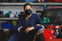 Misto gara femminile meccanico auto indossando maschera viso e tuta, guardando la fotocamera. imprenditore indipendente al garage di assistenza auto durante coronavirus covid 19 pandemia. — Foto stock