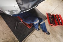 Baixa seção do mecânico do carro feminino vestindo macacão, deitado em uma placa, verificando o carro. proprietário de empresa independente na garagem de manutenção de carro. — Fotografia de Stock