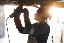 Corrida mista feminino carro mecânico vestindo macacão, usando moedor de ângulo. proprietário de empresa independente na garagem de manutenção de carro. — Fotografia de Stock
