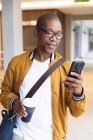 Sonriente negocio afroamericano masculino creativo usando smartphone y sosteniendo café. empresarios creativos independientes que trabajan en una oficina moderna. - foto de stock
