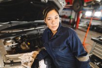 Mixed-Race-Mechanikerin trägt Overalls, inspiziert Auto und blickt in die Kamera. Selbstständiger Unternehmer in der Kfz-Werkstatt. — Stockfoto