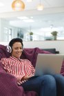 Sonriente mujer caucásica negocio creativo usando auriculares, acostado en el sofá y el uso de la computadora portátil. empresarios creativos independientes que trabajan en una oficina moderna. - foto de stock