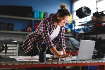 Mixed race female car mechanic standing at desk, using laptop in workshop. independent business owner at car servicing garage. — Stock Photo