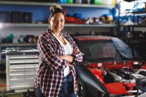 Smiling mixed race female car mechanic standing with crossed hands and looking at camera in workshop. independent business owner at car servicing garage. — Stock Photo