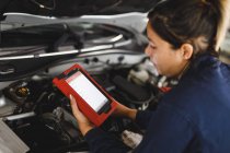 Mixed race female car mechanic wearing overalls, inspecting car and using tablet. independent business owner at car servicing garage. — Stock Photo
