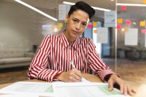 Grave entreprise féminine caucasienne créative assis au bureau, prenant des notes et regardant la caméra. entrepreneurs créatifs indépendants travaillant dans un bureau moderne. — Photo de stock