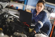 Mixed race female car mechanic wearing overalls, inspecting car, using laptop. independent business owner at car servicing garage. — Stock Photo