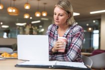 Pensativo negocio femenino caucásico creativo celebración de café y el uso de la computadora portátil en la cafetería del lugar de trabajo. empresarios creativos independientes que trabajan en una oficina moderna. - foto de stock