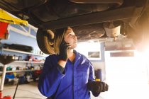 Mécanicien automobile mixte féminin en salopette, inspectant la voiture, parlant par smartphone. propriétaire d'entreprise indépendant au garage d'entretien de voiture. — Photo de stock