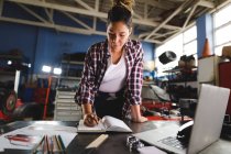 Mixed race female car mechanic sitting at desk, doing paperwork in workshop. independent business owner at car servicing garage. — Stock Photo