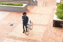 Sonriente negocio femenino afroamericano creativo sosteniendo café y caminando con bicicleta. empresarios creativos independientes que trabajan en una oficina moderna. - foto de stock