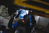 Mixed race female car mechanic wearing overalls, welding car part in workshop. independent business owner at car servicing garage. — Stock Photo