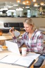 Pensativo negocio femenino caucásico creativo celebración de cookies y el uso de ordenador portátil en la cafetería del lugar de trabajo. empresarios creativos independientes que trabajan en una oficina moderna. - foto de stock