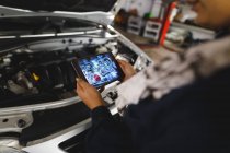 Руки автомеханика смешанной расы носят комбинезон, осматривают автомобиль, используют планшет. независимый владелец автосервиса в гараже. — стоковое фото