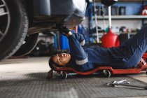 Mécanicien automobile mixte féminin en salopette, allongé sur une planche, vérifiant la voiture. propriétaire d'entreprise indépendant au garage d'entretien de voiture. — Photo de stock
