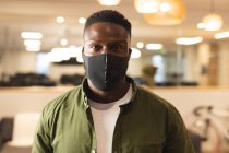 Portrait d'un homme afro-américain créatif portant un masque facial au travail, regardant vers la caméra. travailler dans les affaires créatives dans un bureau moderne pendant la pandémie de coronavirus. — Photo de stock