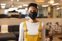 Porträt einer kreativen Frau gemischter Rasse mit Gesichtsmaske bei der Arbeit, die in die Kamera blickt. Arbeit im kreativen Geschäft in einem modernen Büro während der Coronavirus-Pandemie. — Stockfoto