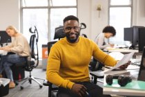 Porträt eines lächelnden afrikanisch-amerikanischen männlichen Kreativen bei der Arbeit, am Schreibtisch sitzend und in die Kamera blickend. Arbeit im kreativen Geschäft in einem modernen Büro. — Stockfoto