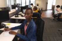 Porträt einer lächelnden afrikanisch-amerikanischen Kreativen bei der Arbeit, am Schreibtisch sitzend, in die Kamera blickend. Arbeit im kreativen Geschäft in einem modernen Büro. — Stockfoto